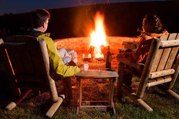 Couple sitting around a campfire in wooden rocking chairs while drinking wine