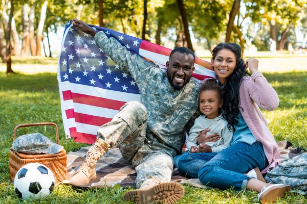 Man wearing a military uniform sitting with his wife and daughter on a picnic blanket and holding up the American flag behind them