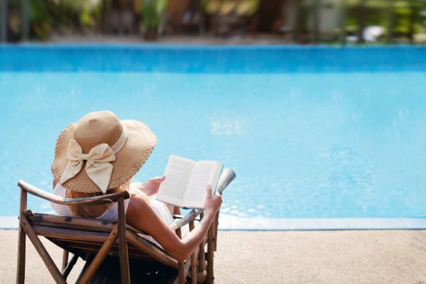 Woman wearing an oversized beach hat reading a book in a lounge chair by a sparkling blue pool